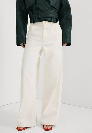 Pantalon blanc à jambes larges et taille haute associé à une veste en cuir noire raccourcie et des sandales rouges à bout carré.