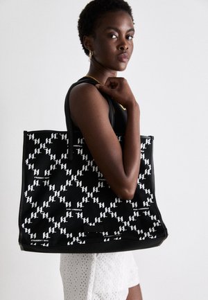 Kvinde med kort hår bærer stor sort taske med hvidt geometrisk mønster og teksten "Karl Lagerfeld", iført hvide shorts med hulmønster.