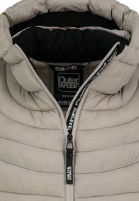 Hellgraue Steppjacke mit hohem Kragen, horizontaler Steppstruktur, schwarzem Innenfutter und einem schwarzen Reißverschluss mit Logodetail.