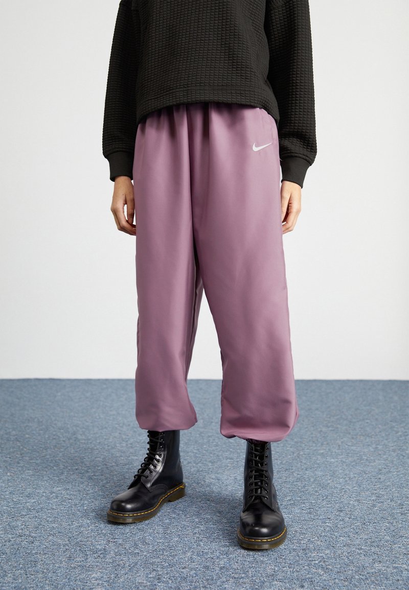 Person klädd i lösa, mauve Nike sweatpants, svarta snörstövlar och en svart strukturerad långärmad topp som står på en blå matta.