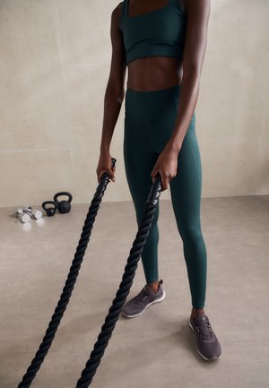 Haut de sport vert et leggings assortis, tenant des cordes de combat noires. Des chaussures de sport grises et des poids sont visibles en arrière-plan.