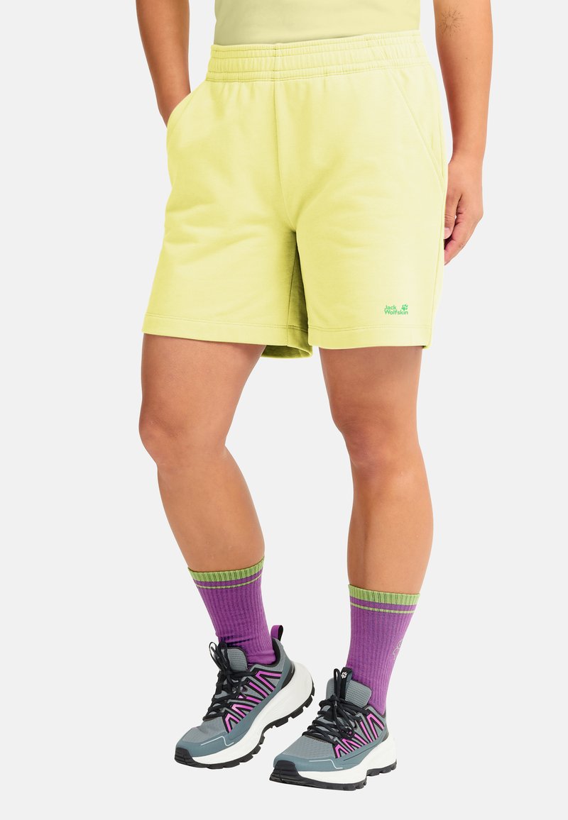 Hellgelbe Baumwollshorts mit elastischem Bund, Seitentaschen und einem kleinen grünen Logo. Kombiniert mit lila und grün gestreiften Socken und grauen Turnschuhen.