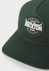 Cappellino da baseball verde scuro con visiera piatta, decorato con un patch circolare ricamato etichettato "Brixton" e motivi di corna in crema e marrone.