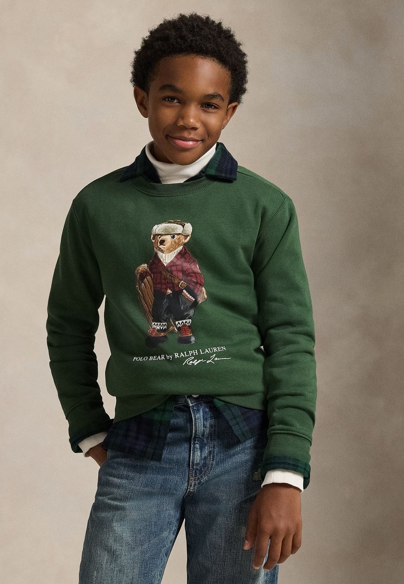 Grön sweatshirt med en björnillustration i rutigt plagg, kombinerad med en blå och grön rutig skjorta under. Bärs med ljusa jeans.