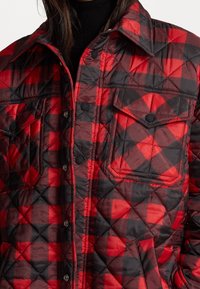 Giacca trapuntata in plaid rosso e nero, con bottoni a pressione, due tasche sul petto e un colletto classico. Tessuto morbido e testurizzato.