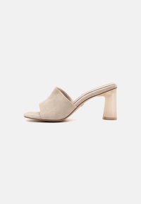Suede slide sandal i beige, med åpen tå, kryssdesign på stroppene, og en gjennomsiktig blokkheel for økt høyde og stabilitet.