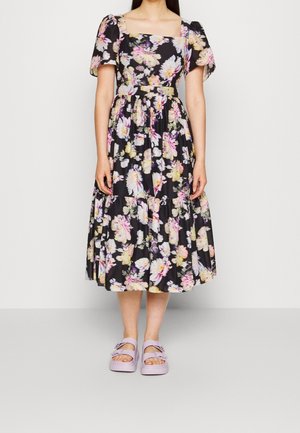 Woman wearing a black floral midi dress with puff sleeves and lilac platform sandals, standing against a plain light background.