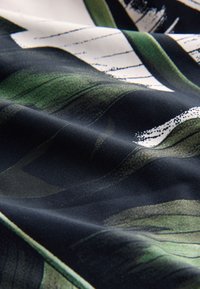 Gros plan d'un tissu sombre avec des motifs abstraits de feuilles vertes et blanches et des plis visibles créant de la texture.