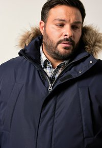 Man som bär en marinblå vinterjacka med pälsbeklädd huva över en svartvit rutiga skjorta, ser nedåt och åt sidan.