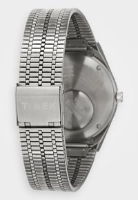 Bracelet de montre Timex en métal argenté avec fermoir, présentant un design de maillons flexibles et un dos de montre visible avec un bouton de réglage.