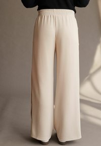 Beige brede benbukser med en smock-elt, lavet af glat stof. Har sidelommer og en afslappet pasform, der strækker sig til gulvet.