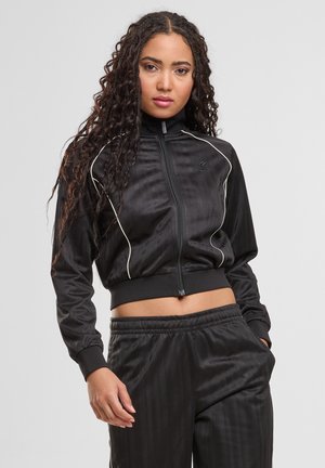 Frau mit langen lockigen Haaren, die eine schwarze, kurz geschnittene Trainingsjacke mit Reißverschluss und weißen Paspeln sowie passende schwarze Trainingshose trägt.