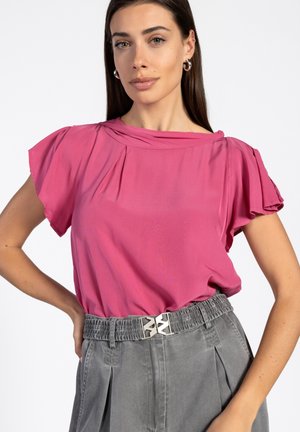 Roze blouse met gedrapeerde halslijn en korte fladdermouwen, gecombineerd met grijze geplooide broek en een decoratieve metalen riemdetail.