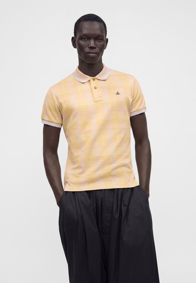 Homme portant un polo jaune clair à manches courtes avec un petit logo et un pantalon noir ample, debout les mains dans les poches.