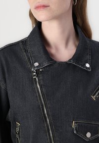 Denimjakke i mørkegrått med høyt krage, sølvglidelås og kontrastfargede gule sømmer. Har glidelås- og knappede lommer.