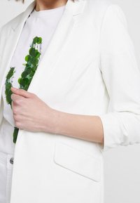 White tailored blazer with a notch lapel, single button closure, and side pocket; worn over a t-shirt with a green cactus graphic.
