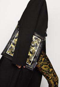Schwarzer Mantel mit schwarzer und goldener gemusterter Versace Jeans Couture Tasche und passenden ornamentalen gold-schwarzen Hosen.