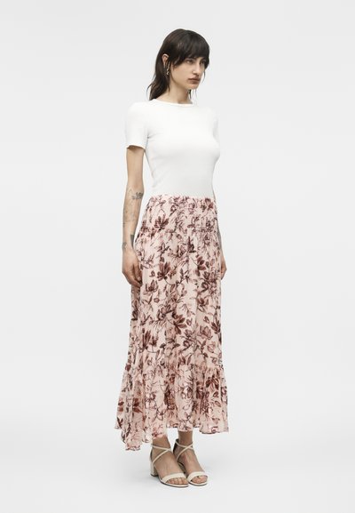 Kvinde med mørkt hår iført en hvid kortærmet top, lang nederdel med pink blomsterprint og beige hælede sandaler, stående i profil.