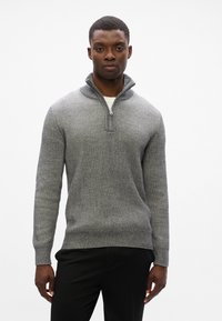 Suéter gris acanalado con un cuello con cremallera, que presenta una tela texturizada y mangas largas, combinado con pantalones oscuros.