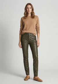 Vrouw die een camelkleurige trui, olijfgroene leren broek en beige loafers draagt, staand tegen een effen lichtgrijze achtergrond.