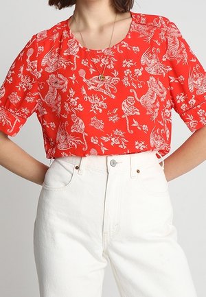 Kobieta w jasnoczerwonej bluzce z białym nadrukiem zwierzęcym i kwiatowym, wpuszczonej w wysokie, białe spodnie.