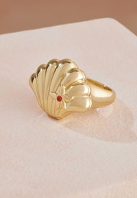 Zlati prstan v obliki školjke z teksturiranimi rebri in majhnim rdečim kamnom kot poudarkom. Gladek, poliran videz z zaobljeno obrobo.