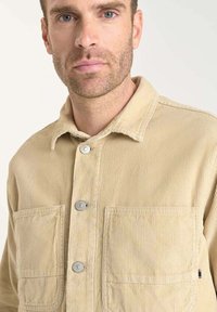 Beige corduroy jas met een kraag, twee voorzakken en metalen drukknoopsluitingen. Ribbelstructuur en gestikte accenten voegen detail toe.