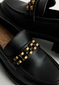 Mocassini in pelle nera con una fascia texturizzata, dettagli in ottone dorato e design intrecciato, punta arrotondata e cuciture visibili.