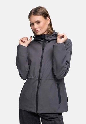 Žena si upravuje ťahadlá na sivom kapucňovom bundu so zipsom, oblečená v tmavých nohaviciach na jednolatom bielom pozadí.