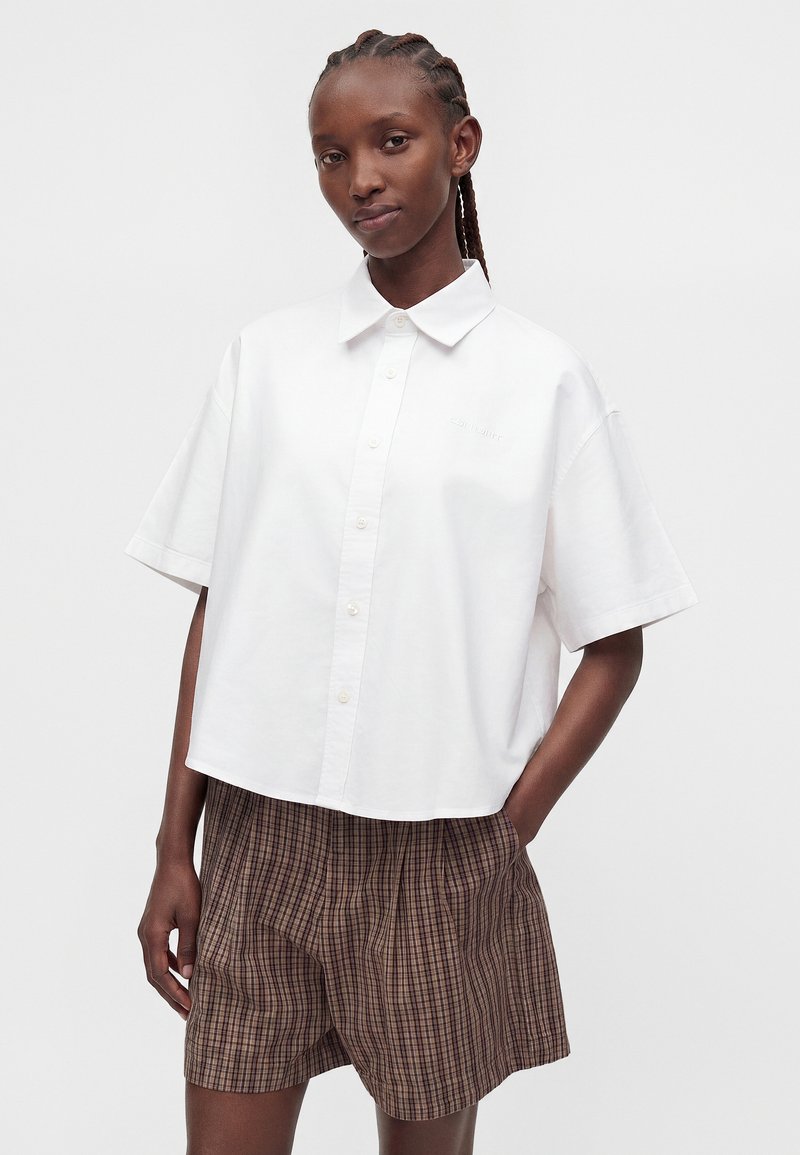 Woman with braided hair wearing a loose white short-sleeve button-up shirt and brown plaid shorts, standing with one hand in pocket.
