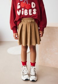 Sweat-shirt rouge avec le texte « GOOD VIBES », jupe plissée en velours côtelé beige, chaussettes rouges à volants et baskets blanches avec des accents imprimé léopard.