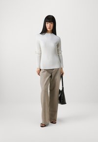 Weißer gerippter Pullover mit langen Ärmeln, kombiniert mit lockeren beigen Jeans. Das Modell hält eine schwarze Handtasche und trägt schwarze Sandalen.