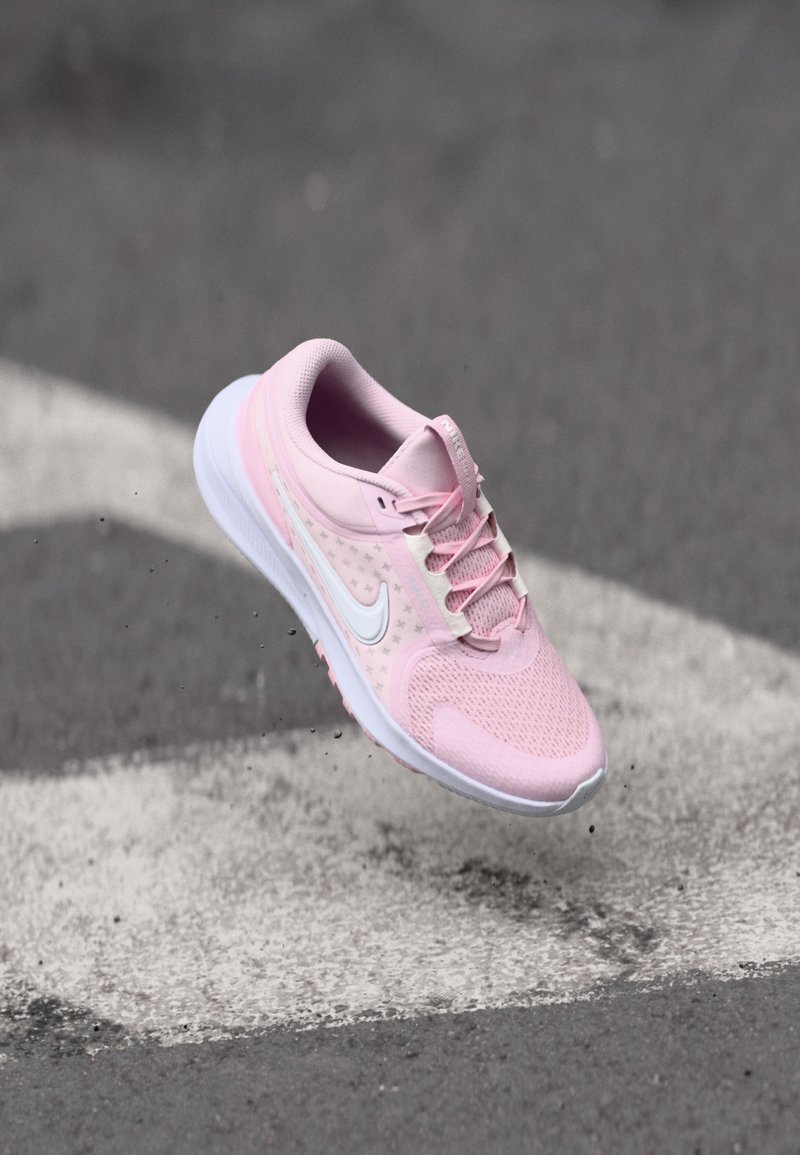 Bottine de course Nike rose en l'air au-dessus d'une surface d'asphalte texturée avec des flèches peintes en blanc.