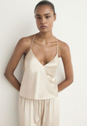 Donna che indossa un top in raso color crema e pantaloni larghi abbinati, in piedi davanti a uno sfondo bianco semplice con le mani dietro la schiena.