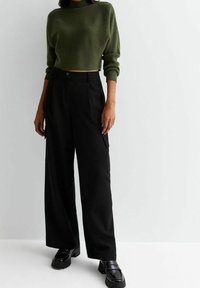 Oliivinvihreä lyhyt neulepusero, jossa resorikuviointi, yhdistetty mustiin, korkeavyötäröisiin leveälahkeisiin housuihin ja kiiltäviin mustiin loafer-kenkiin.