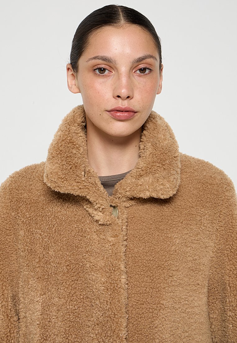Femme aux cheveux tirés en arrière portant un manteau teddy fluffy de couleur brun clair avec un large col et un devant boutonné.