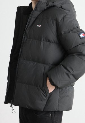 Person trägt eine schwarze Steppjacke mit Kapuze, Hand in der Tasche, zeigt die Tommy Hilfiger-Logos auf Brust und Ärmel.