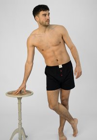Boxer en coton noir avec des accents rouge sur les boutons et une ceinture élastique, présentés sur un modèle à côté d'une table d'appoint en bois clair.