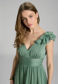 Kleid aus weichem grünen Stoff mit tiefem V-Ausschnitt, gerafftem Oberteil und Rüsche an der Schulter. Fließender Rock mit glatter Textur.