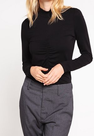 Vrouw in een nauwsluitend zwart top met lange mouwen en een geplooide voorkant, gecombineerd met hooggetailleerde, grijze, op maat gemaakte broek, tegen een effen achtergrond.