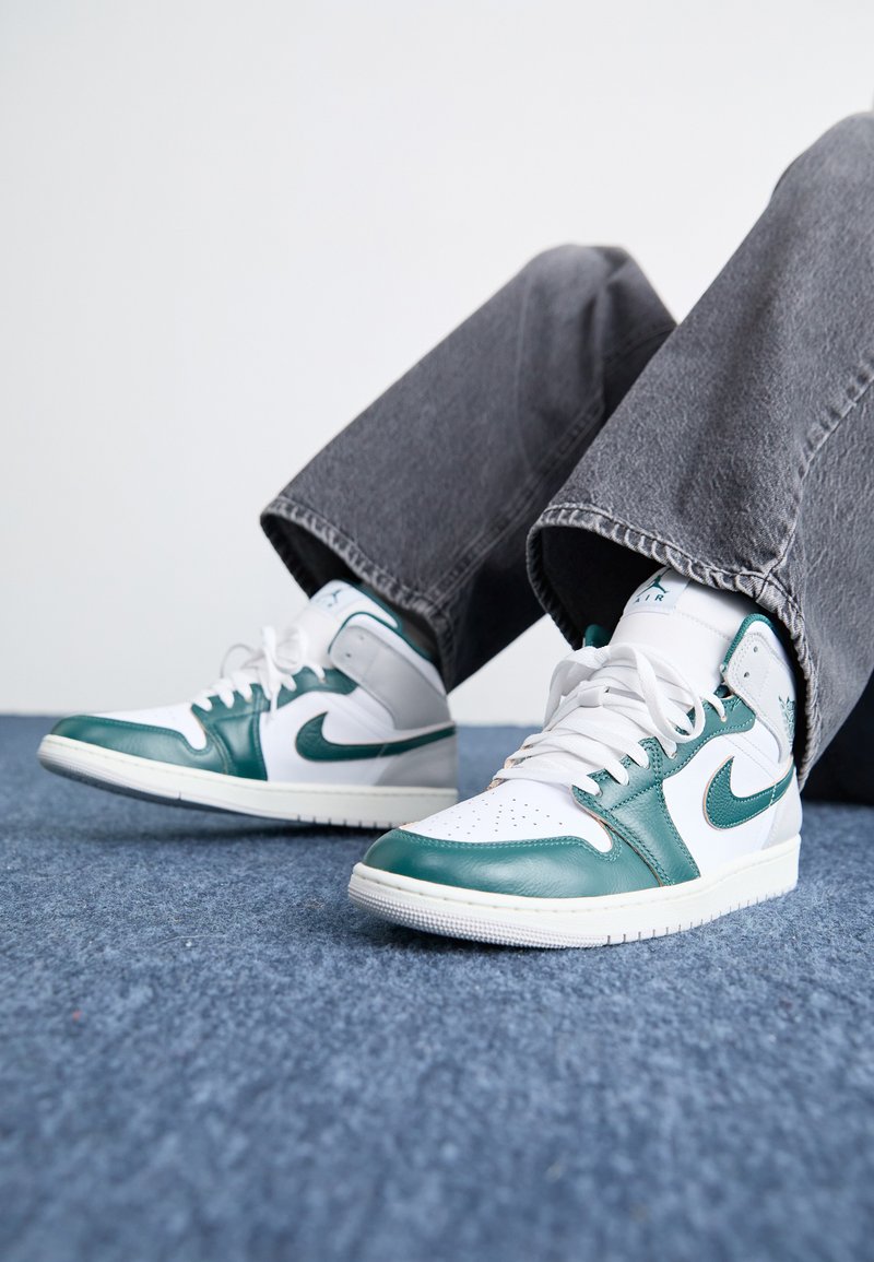 Adidași înalți din piele albă și teal, cu vârf perforat, șireturi albe și o talpă texturată, asortați cu pantaloni largi gri.