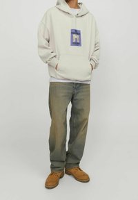 Felpa grigia chiara con cappuccio, tasca frontale e design grafico. Abbinata a jeans blu larghi e stivali da lavoro marroni. Outfit casual.