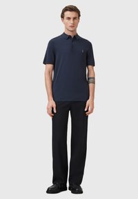 Marineblauwe poloshirt met korte mouwen, knoopsluiting en een klein logo op de borst, gecombineerd met zwarte broeken en schoenen.