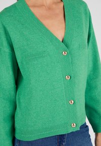Groene cardigan met een V-hals ontwerp, twee voorzakken en gouden knopen. De gladde textuur en de ribgebreide details aan de onderkant verbeteren de algehele uitstraling.