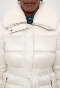 Puffer coat in cream with a fluffy white collar, belted waist, and quilted design. Features silver hardware and zippered pockets.