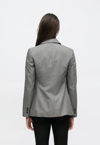 Blazer gris sur mesure avec col cranté, manches longues, coupe structurée et poignets boutonnés. Confectionné dans un tissu texturé, sans motifs visibles.