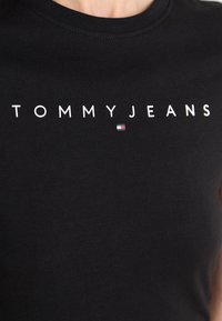Černé bavlněné tričko s potiskem "TOMMYJEANS" v bílé barvě. Obsahuje malé logo v červené, bílé a modré barvě poblíž textu.