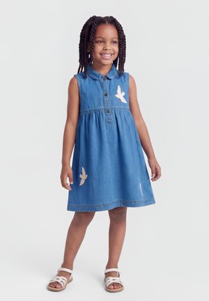 Lachend meisje met gevlochten haar, gekleed in een mouwloze spijkerjurk met vogelapplicaties en witte sandalen, staand tegen een effen achtergrond.