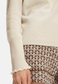Cremfarbener Rippstrickpullover mit ausgestellten Bündchen, kombiniert mit braunen, strukturierten Shorts, die ein gewebtes Muster sowie Akzente in Weiß und Rostrot aufweisen.