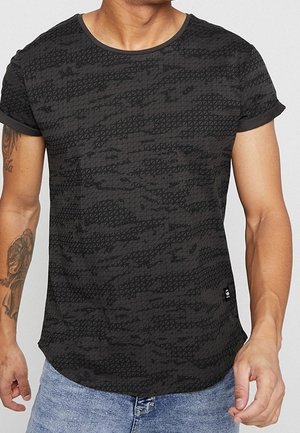 Homme portant un t-shirt manches courtes à motifs sombres avec les manches retroussées et un jean bleu clair, montrant des tatouages sur le bras gauche.